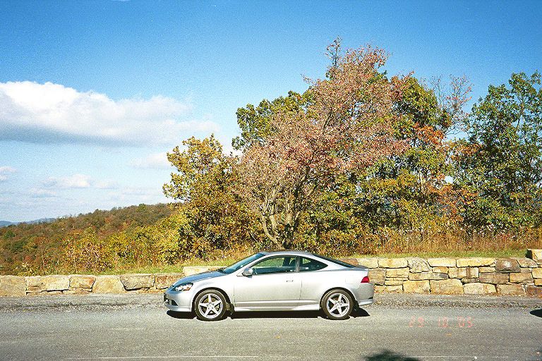 Silver coupe parked in side profile by a stone wall with fall trees behind