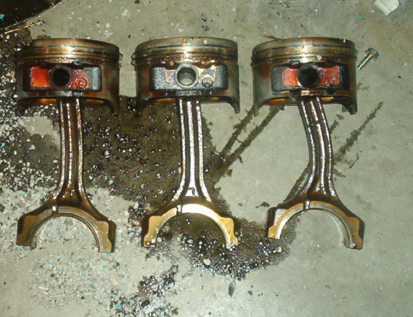Three greasy pistons with connecting rods attached, lying on a concrete floor