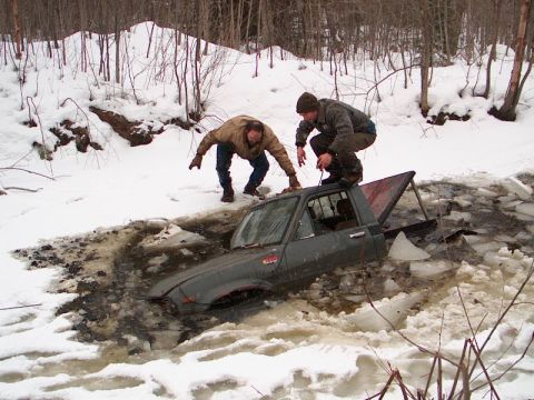 1995 Ford Bronco - Two individuals observe a dark pickup truck stuck in a frozen body of water surrounded by snow-snowy woods.
