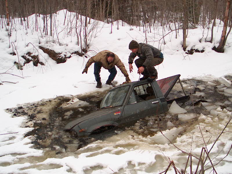 Dark pickup truck partially submerged in icy water with two people on top