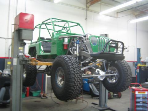 1990 Ford Bronco - green tube-chassis off-road buggy raised on a two-post lift in a shop, showing its suspension and large mud-terrain tires.