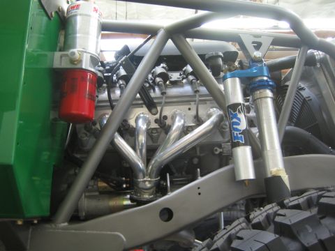 1990 Ford Bronco - Detail of a green off-road buggy showing polished exhaust headers, an oil filter, tube chassis, and a blue Fox coilover shock.