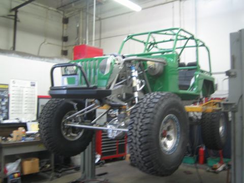 1990 Ford Bronco - green tube-chassis off-road buggy raised on a two-post lift, viewed from the front showing suspension and large tires.