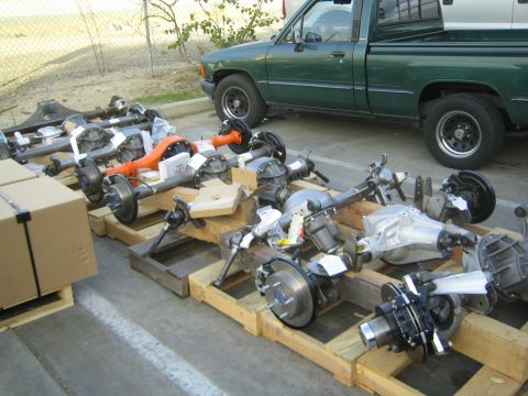 1990 Ford Bronco - Several assembled vehicle axles staged on wooden pallets in a yard with a dark green pickup truck parked behind.
