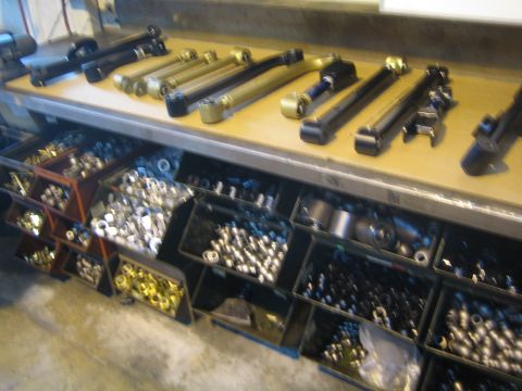 1990 Ford Bronco - Fabricated suspension control arms laid out on a workbench above open bins full of bushings, bolts, and hardware.