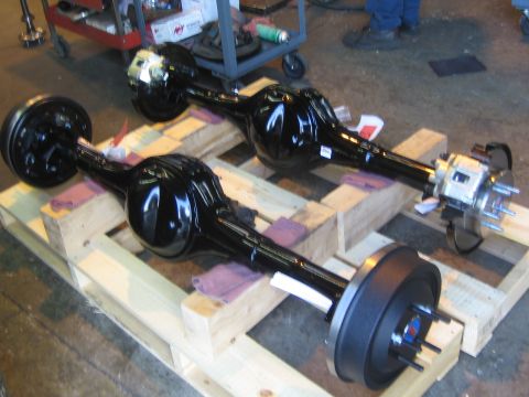 1990 Ford Bronco - Two finished gloss-black rear axle assemblies with brake drums and hubs sit on a wooden pallet in a garage.