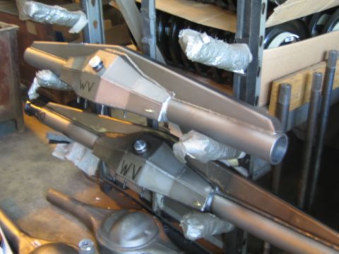 1990 Ford Bronco - Fabricated raw steel axle housings hand-marked WV are stacked on a metal warehouse storage rack.