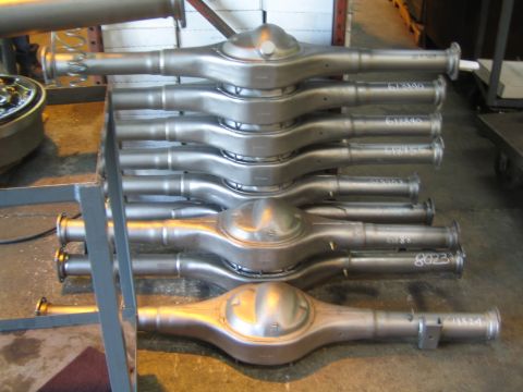 1990 Ford Bronco - Machined steel axle housings, several with handwritten part numbers, are stacked on a metal shop rack.