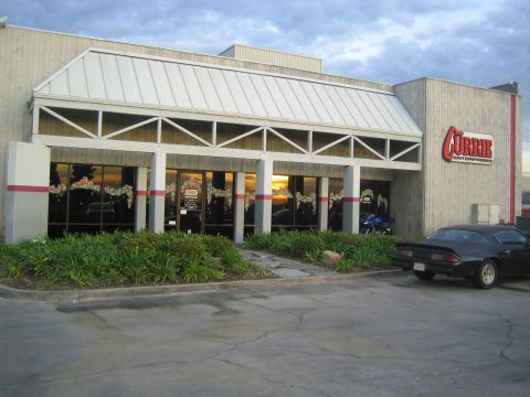 1990 Ford Bronco - Currie Enterprises building at dusk with holiday-decorated windows and a black coupe parked in front.
