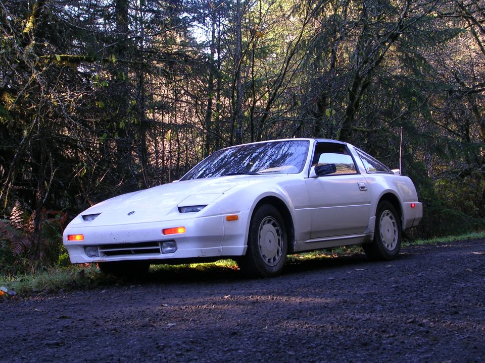 White Nissan 300ZX Z31 coupe on gravel road in evergreen forest, 3/4 front view