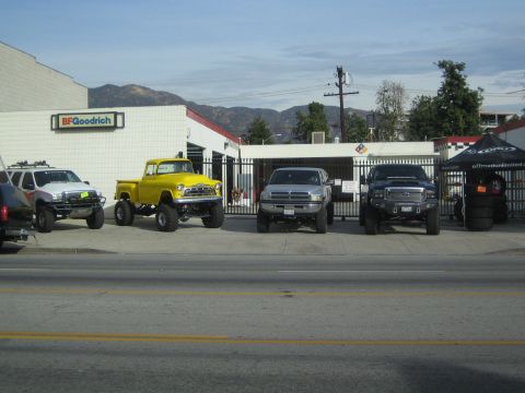 1990 Ford Bronco - Four lifted trucks, including a yellow vintage pickup, line up outside a BFGoodrich shop with mountains behind.