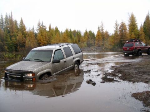 2000 Chevrolet Tahoe - A silver SUV is stuck in a muddy puddle while a red Jeep attempts to pull it free.