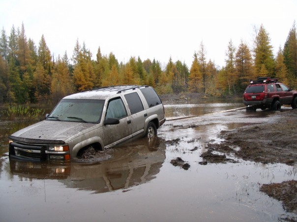 Muddy tan Chevy SUV stuck in a flooded mud hole with red Jeep recovering it