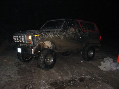 1982 Ford Bronco - A muddy 1980s SUV is parked on an unpaved surface at night, showing its lifted suspension and off-road tires.