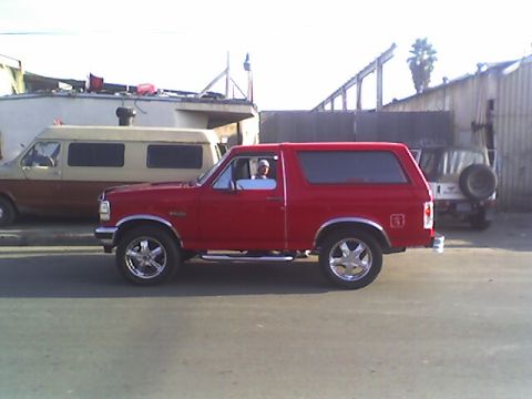 1990 Ford Bronco - red two-door full-size SUV with chrome wheels and a raised stance is parked in profile near shop buildings.