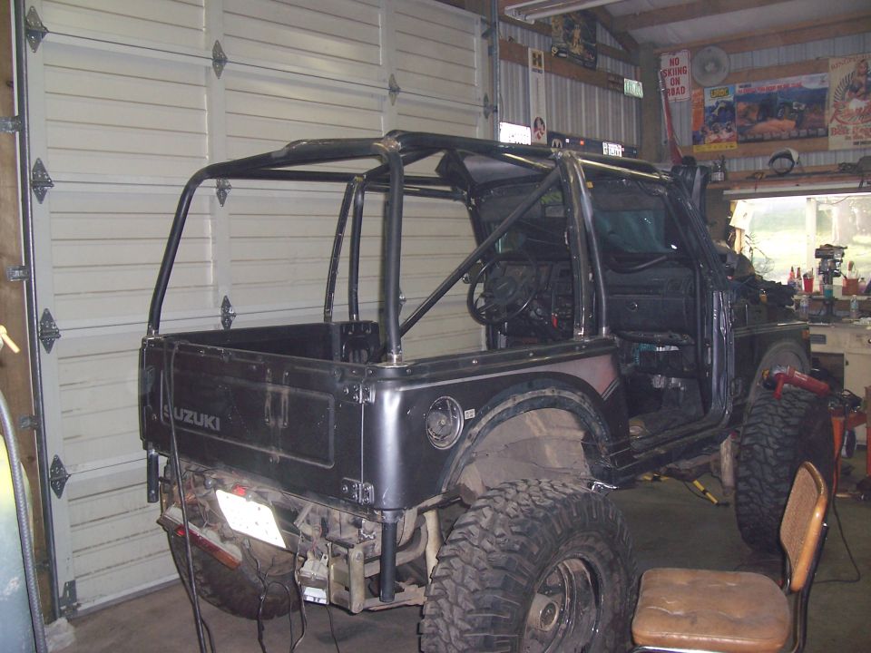 Black Suzuki Samurai with full roll cage, no doors or top, on big mud tires inside a garage, rear view