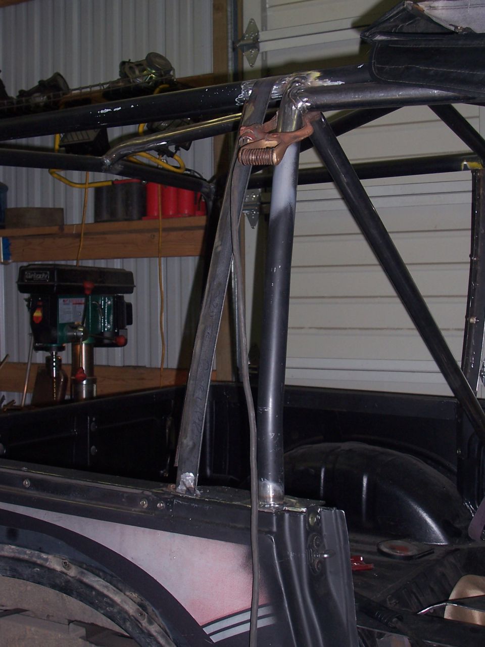 Roll cage tubing welded to black Suzuki Samurai tub in garage, welding ground clamp attached to bar