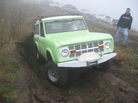 1999 Jeep Grand Cherokee - A vintage Ford Bronco navigates a muddy off-road course, with several other vehicles visible on the hill behind it is climbing.
