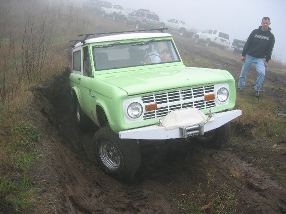 Green 1970s Ford Bronco with white roof and bumper, on a muddy incline, with a man standing nearby and other SUVs in the background