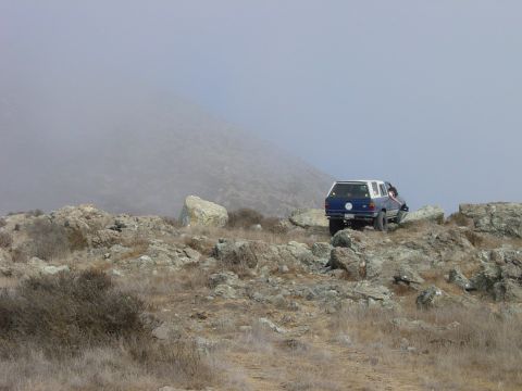 1999 Jeep Grand Cherokee - A blue and white SUV navigates a rocky, off-road landscape under a foggy sky.