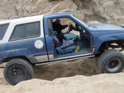1999 Jeep Grand Cherokee - A blue pickup truck with a camper shell is stuck in a sandy ditch, with a person in the driver's seat.