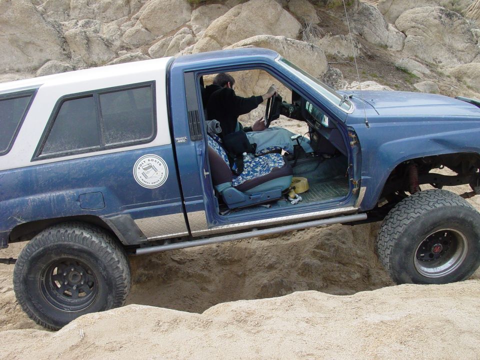 Blue pickup truck with camper shell stuck in sand, driver visible inside