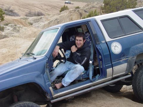 1999 Jeep Grand Cherokee - Man sits in the driver's seat of a modified blue pickup truck on an off-road trail.