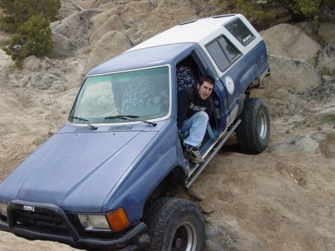 1999 Jeep Grand Cherokee - A man sits in the open door of a blue pickup truck with a white camper shell, navigating a rocky, off-road path.