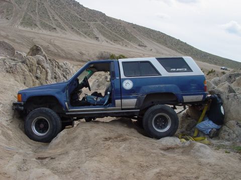 1999 Jeep Grand Cherokee - A blue pickup truck with a white camper shell navigates a rocky off-road course, with a person preparing a tow strap.