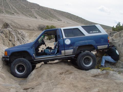 1999 Jeep Grand Cherokee - A blue SUV with a white camper shell is shown navigating a rocky, off-road environment, with text on the camper reading 'MT. STATES'.