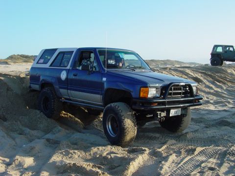 1999 Jeep Grand Cherokee - A blue pickup truck with a camper shell navigates sandy terrain, with another vehicle visible in the background.