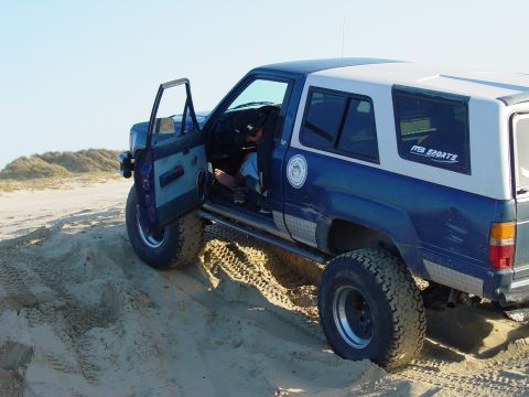 1999 Jeep Grand Cherokee - This blue and white SUV is shown off-road on a sand dune with its driver's door open. A decal reads "MB SPORTS".