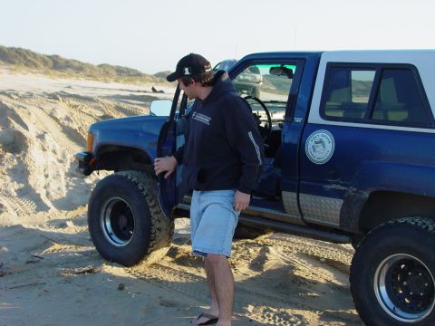 1999 Jeep Grand Cherokee - A man opens the door of a modified blue SUV parked on a sandy dune area, with another vehicle in the background.