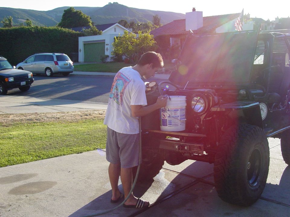 guy working on a dark green Jeep CJ-7 with hood up, bucket and hose nearby