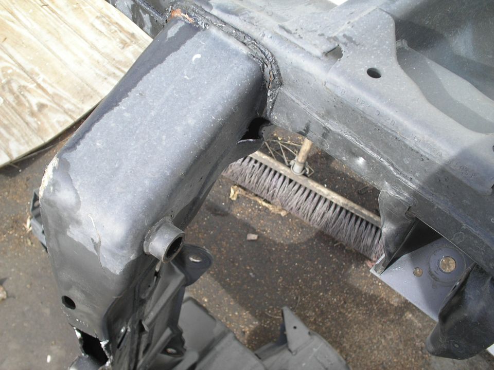 Close-up of a dark, heavy metal vehicle frame part with visible welds and some surface wear. A broom is visible in the background