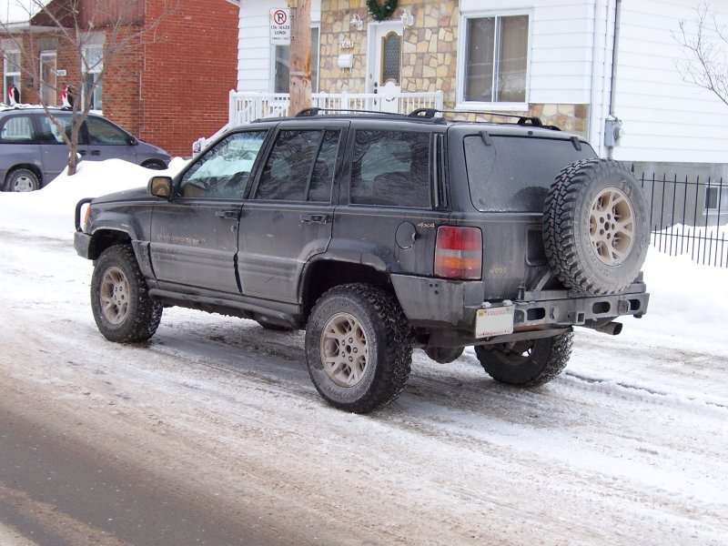 Black lifted Jeep Grand Cherokee, rear 3/4 view, with spare tire carrier, parked on snowy street.