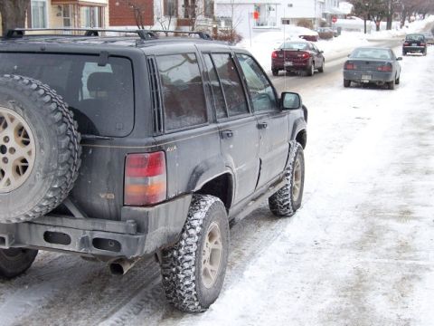 1996 Jeep Grand Cherokee - Rear view of a 1996 Jeep Grand Cherokee on a snow-covered street with other vehicles in the background.