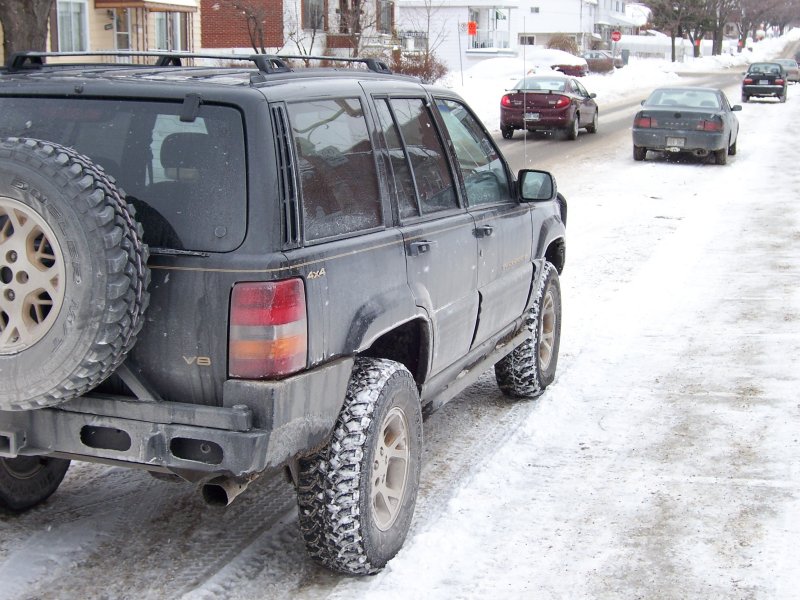 Black Jeep Grand Cherokee with rear tire carrier and mud tires on a snow-covered street