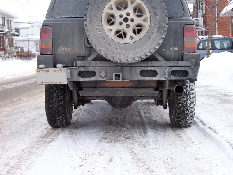 Rear of dark Jeep Grand Cherokee with custom steel bumper and full-size spare tire carrier on a snowy street