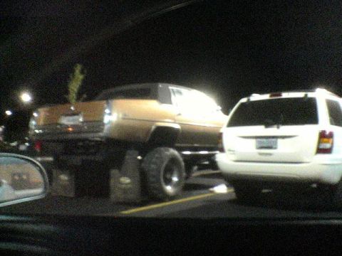 1994 Ford Probe - A modified gold sedan with large off-road tires is parked beside a white SUV.