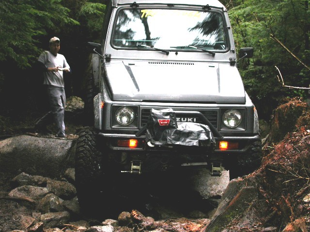 Silver Suzuki Samurai with Warn winch crawling over creek-bed boulders, man standing alongside