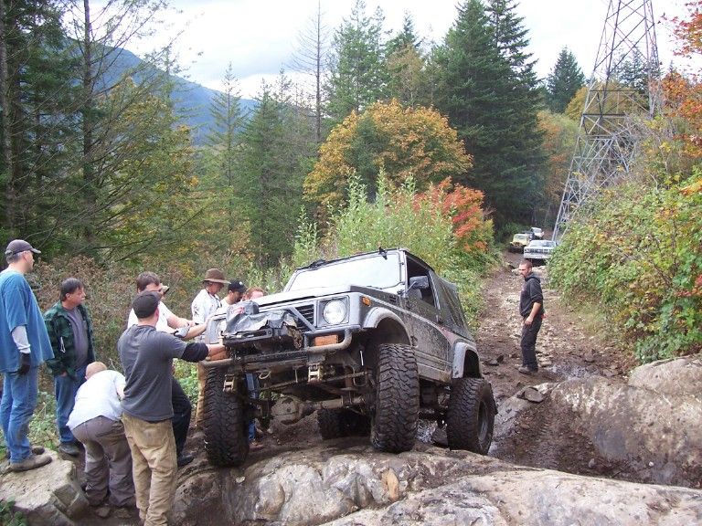 Lifted Suzuki with winch bumper crawling rocks on a trail surrounded by spectators and fall trees