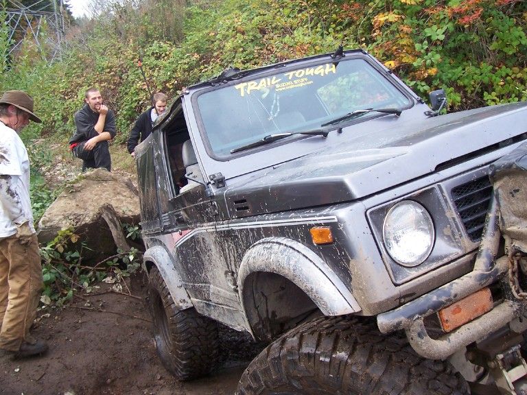 Muddy gray Suzuki Samurai with Trail Tough banner stuck on rocks, men watching from the bank