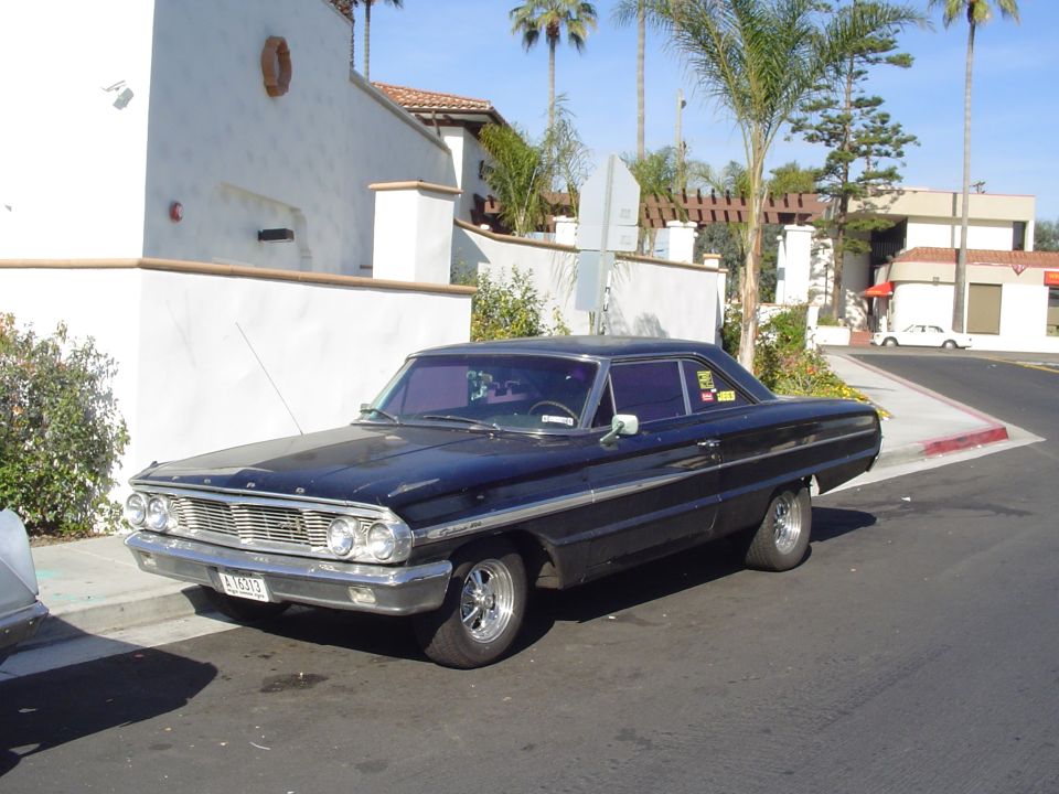 Black 1960s Ford two-door hardtop parked at a curb, front three-quarter view with palm trees