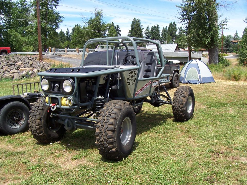 Toyota-front tube-chassis rock buggy on grass, flat hood, dual headlights, big mud tires, tent in background