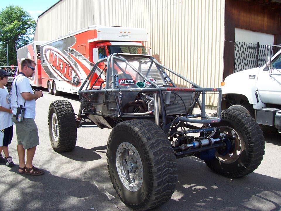 Bare-metal tube-chassis rock buggy on chrome beadlocks parked in a driveway, Maxxis trailer behind