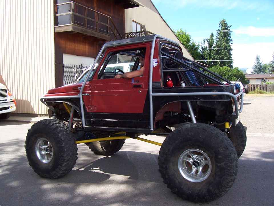 Lifted red Suzuki Sidekick on big mud tires and beadlocks parked beside a building, driver inside