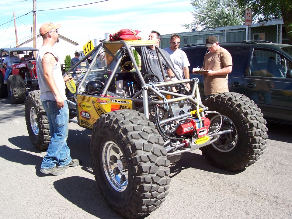 Yellow tube-chassis rock buggy with rear-mounted winch and sponsor decals on pavement, men around it