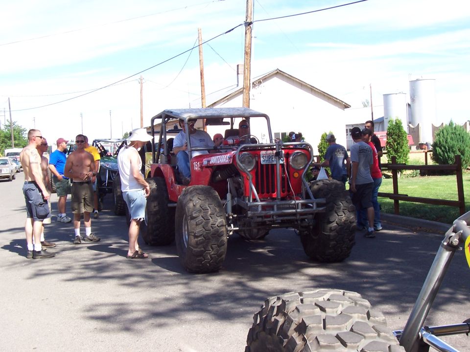 Red flatfender rock buggy with riders driving on a street past a crowd, barn and silos behind