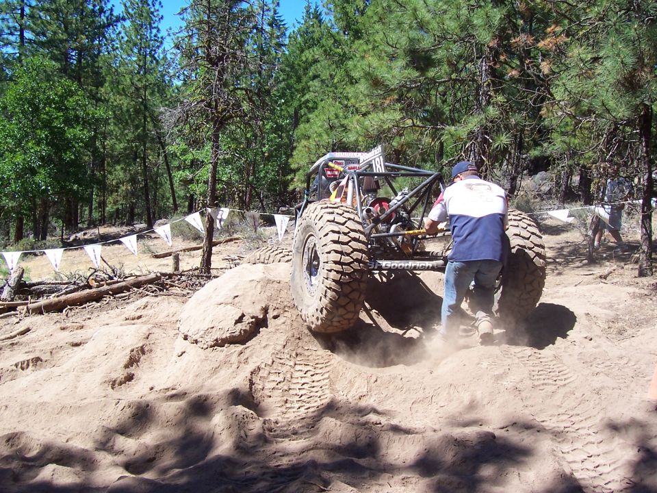 Rear of a tube buggy churning a sandy mound as a spotter pushes it forward